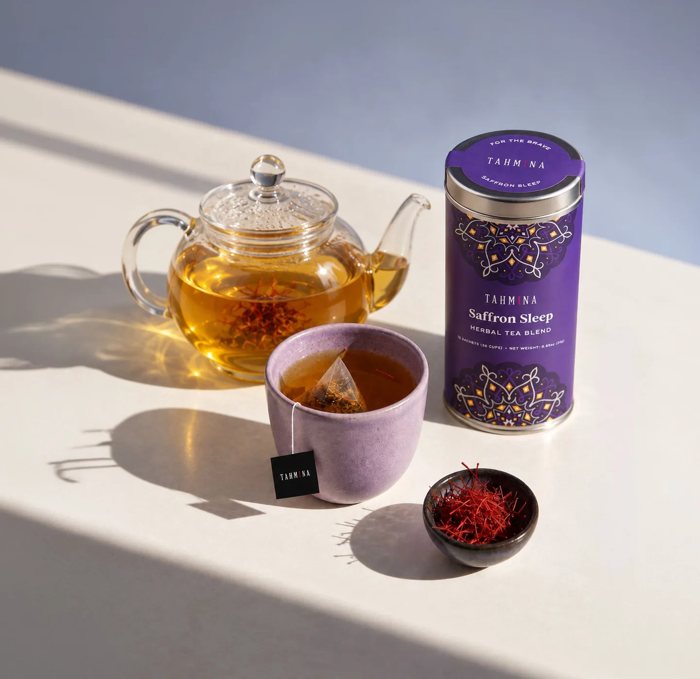 Saffron tea in a glass teapot and a lilac cup with a Tahmina sachet, beside a Saffron Sleep tin and a dish of saffron threads