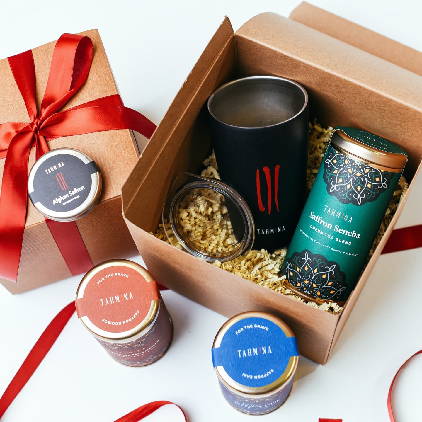 An open kraft gift box holding a black Tahmina tumbler and a Saffron Sencha tin, beside a red-ribboned box topped with Afghan Saffron and tins of Saffron Rooibos and Saffron Chai